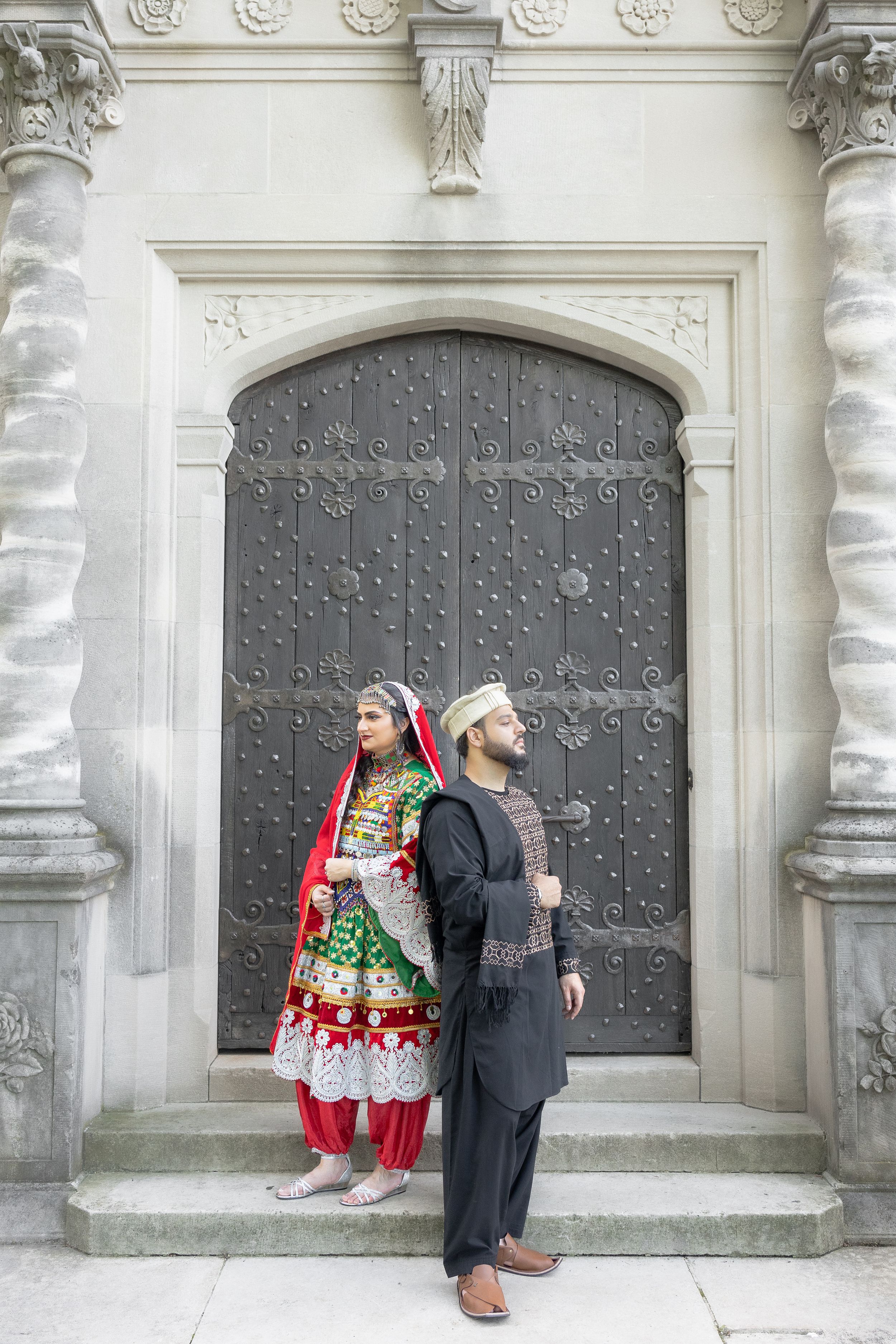 A couple in traditional South Asian attire stands against an ornate gray door with decorative stone columns.