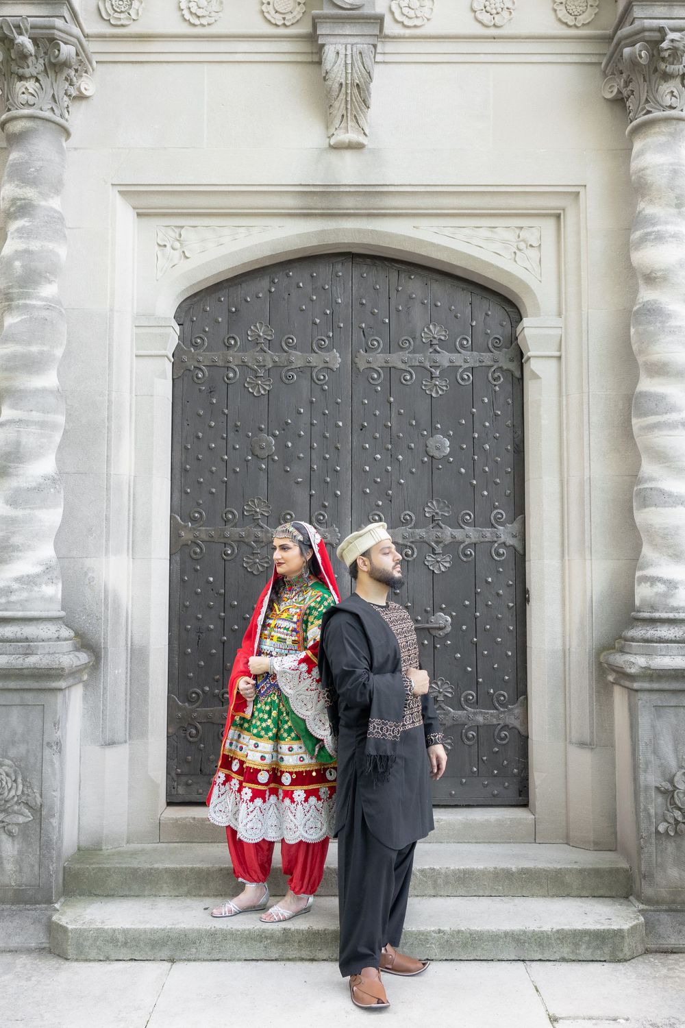 A couple in traditional South Asian attire stands against an ornate gray door with decorative stone columns.