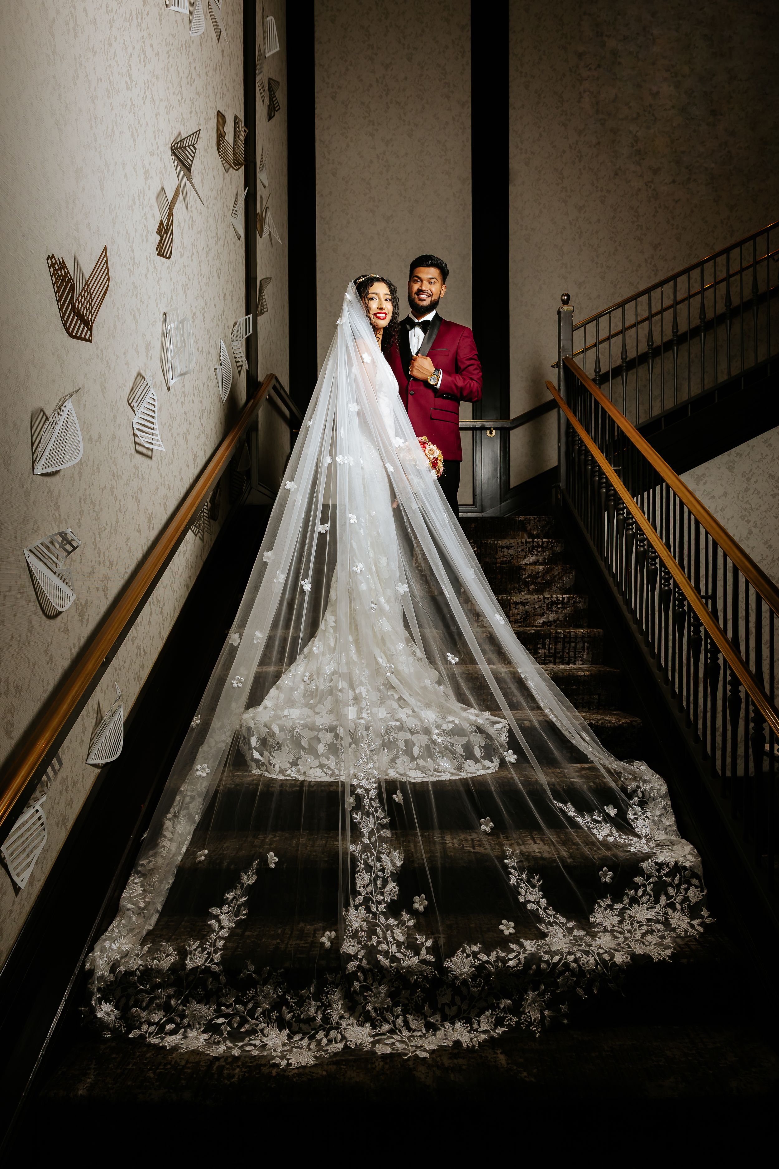 A wedding couple poses on a dramatic staircase with a flowing lace veil creating an elegant silhouette against dark walls.
