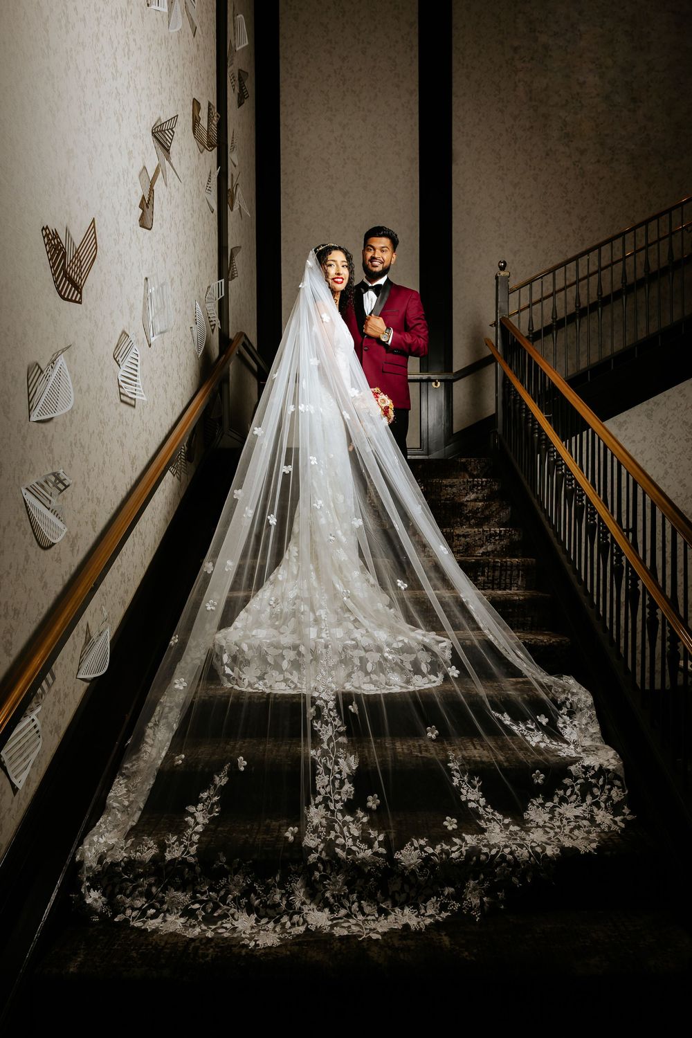 A wedding couple poses on a dramatic staircase with a flowing lace veil creating an elegant silhouette against dark walls.