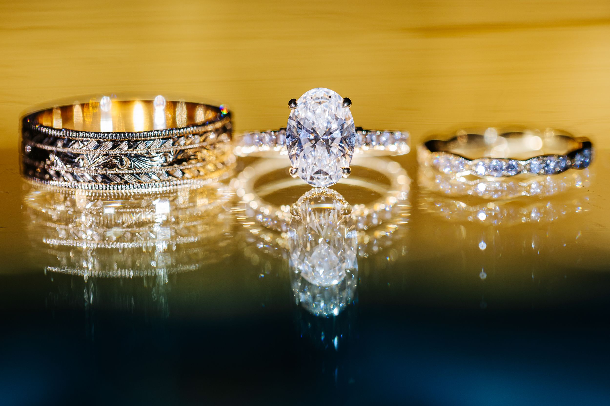 Three sparkling diamond wedding rings with intricate designs displayed on reflective black surface with golden lighting.