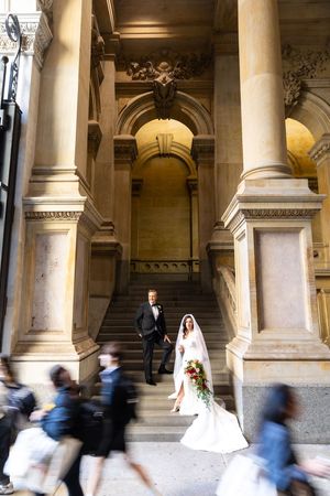 A couple poses on grand stone steps of a historic building while pedestrians walk past in a blur of motion.