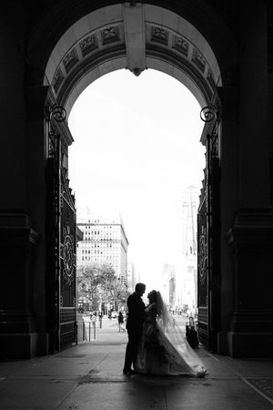 Two silhouettes embrace under an ornate stone archway on a city street captured in dramatic black and white photography.