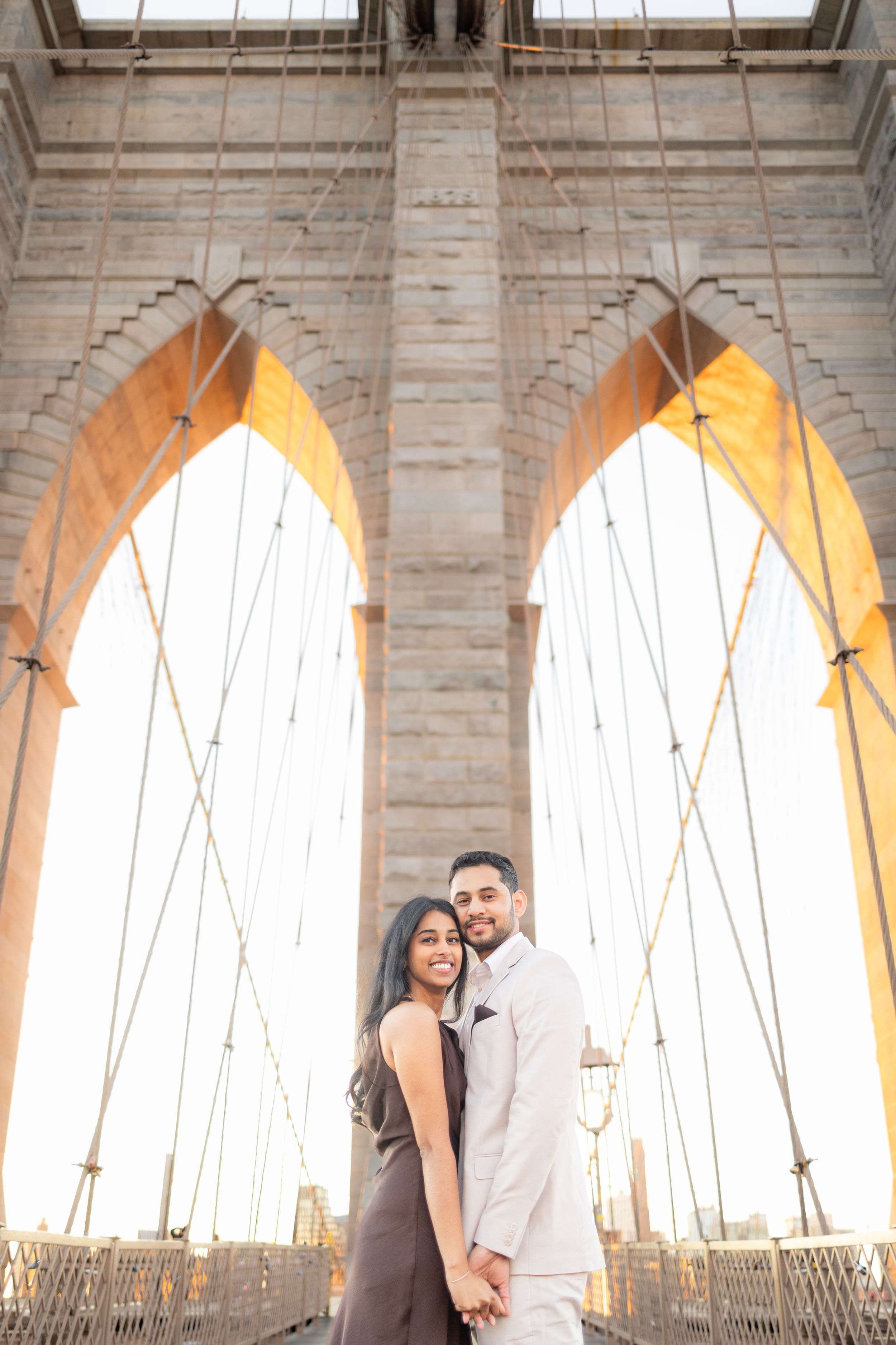 A couple poses beneath the iconic stone arches of the Brooklyn Bridge at sunset.