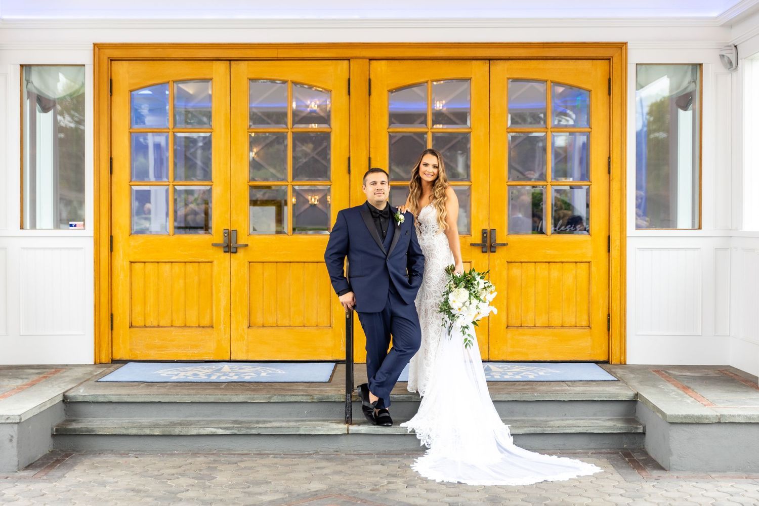 Couple poses in front of bright yellow double doors with glass panes and white building exterior.