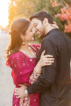 A romantic couple embraces outdoors during golden hour, with her wearing a sparkling red traditional South Asian outfit.
