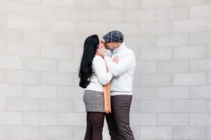 A romantic couple in cozy winter sweaters share a tender moment against a grey concrete wall.