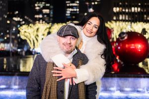 Two people in winter coats smile together at an outdoor ice rink with city lights and holiday decorations in background.