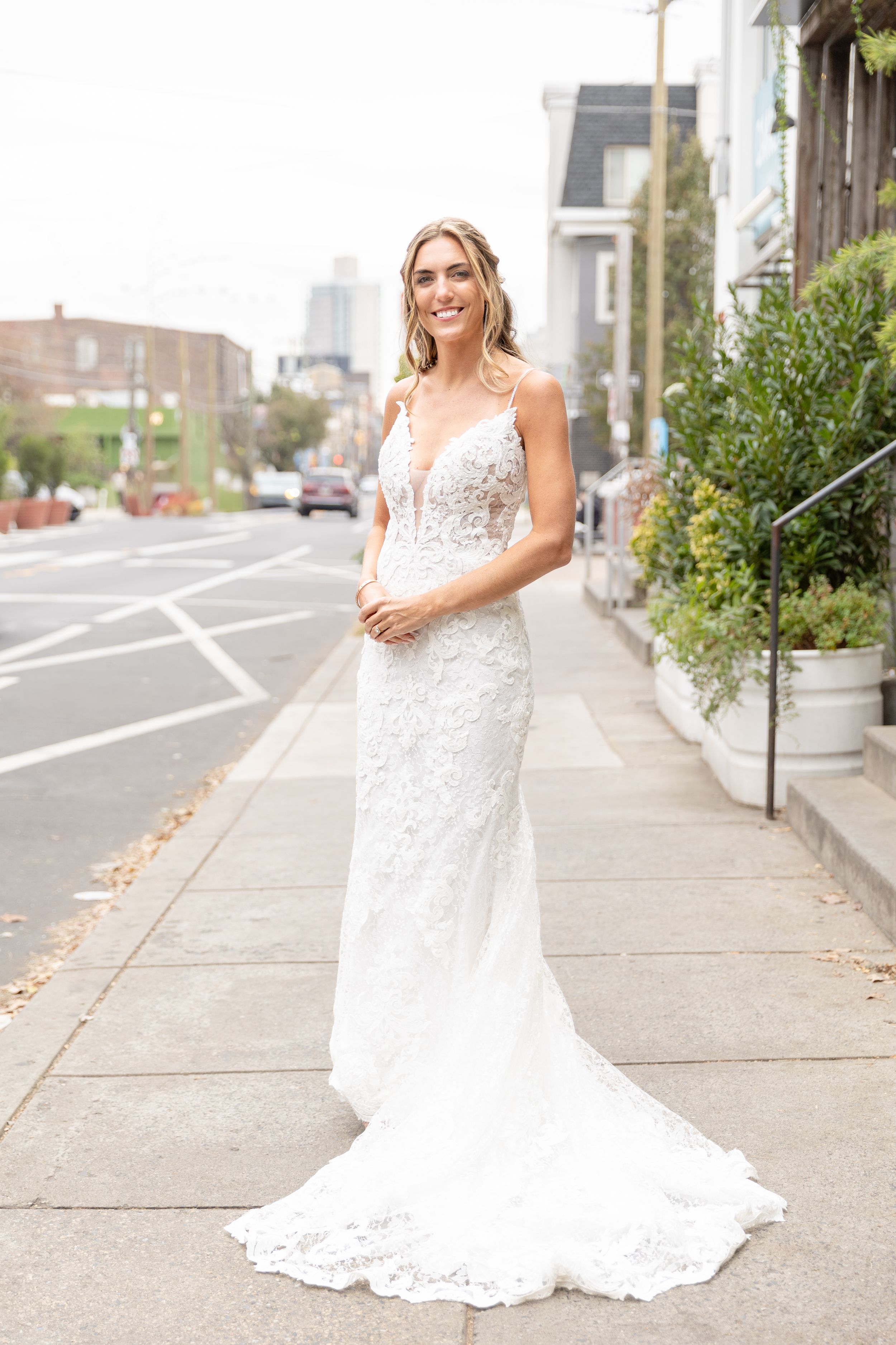 A stunning white lace wedding gown with a plunging neckline and fitted silhouette is showcased on a city sidewalk.