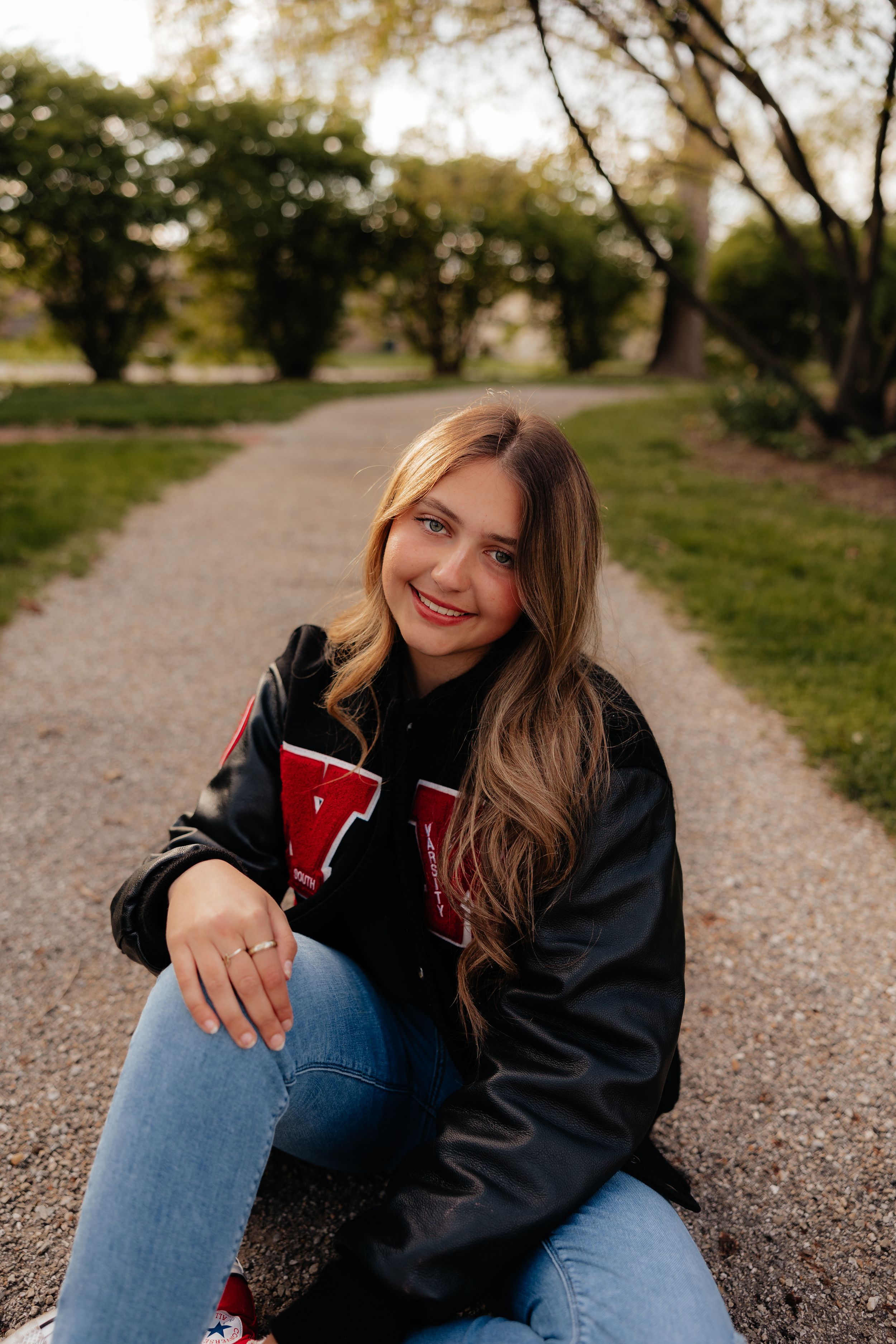 Casual portrait of person in varsity jacket sitting on path outdoors.