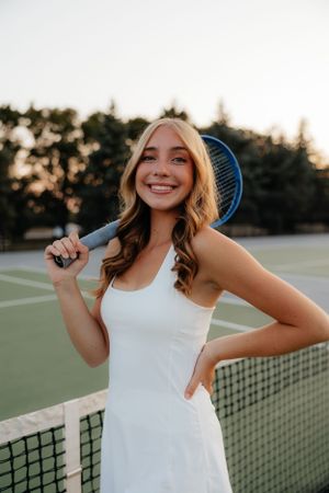 Tennis player in white dress smiling with racquet on court at sunset.
