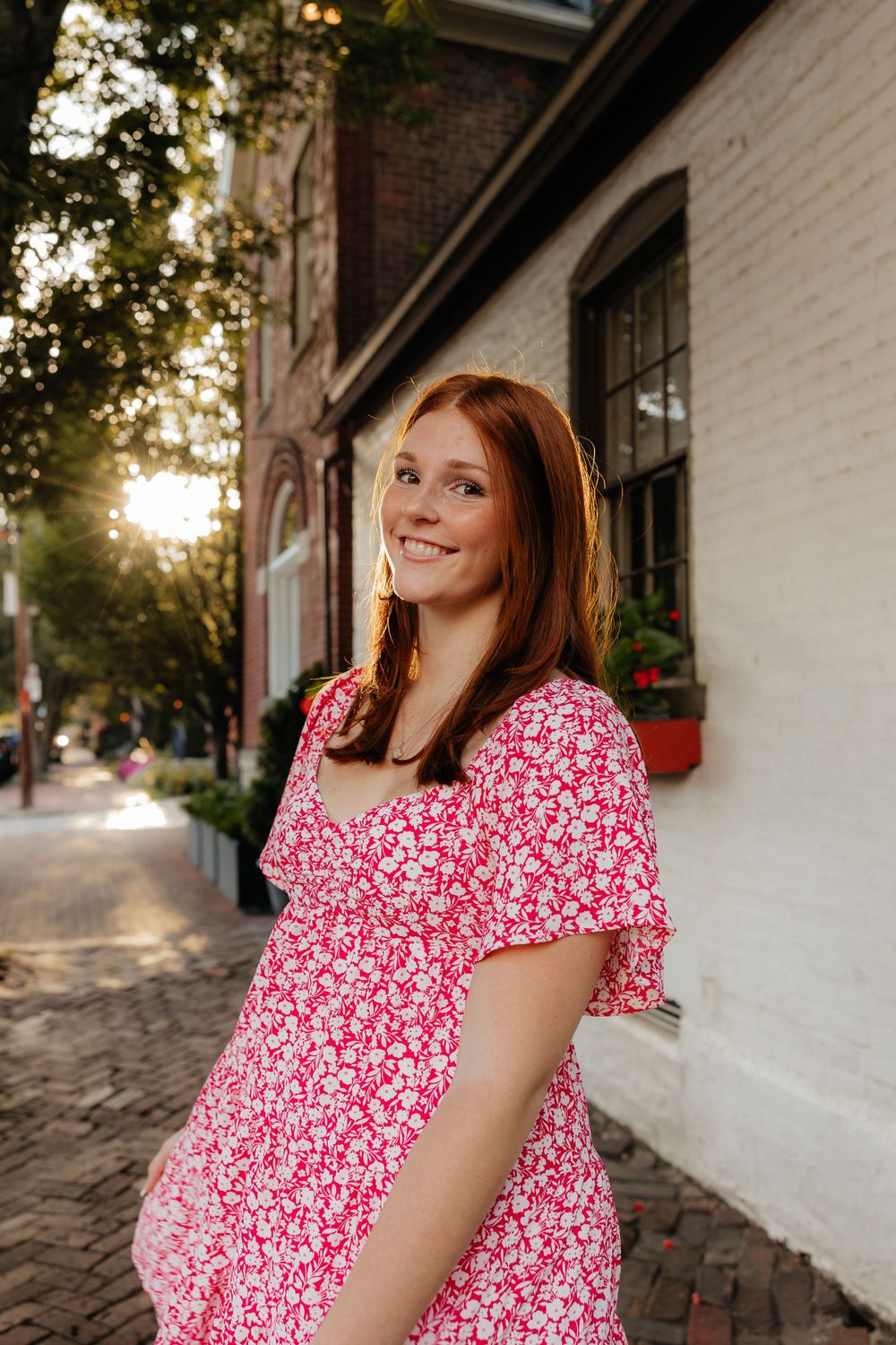 Woman in red floral dress poses along historic brick building sidewalk.