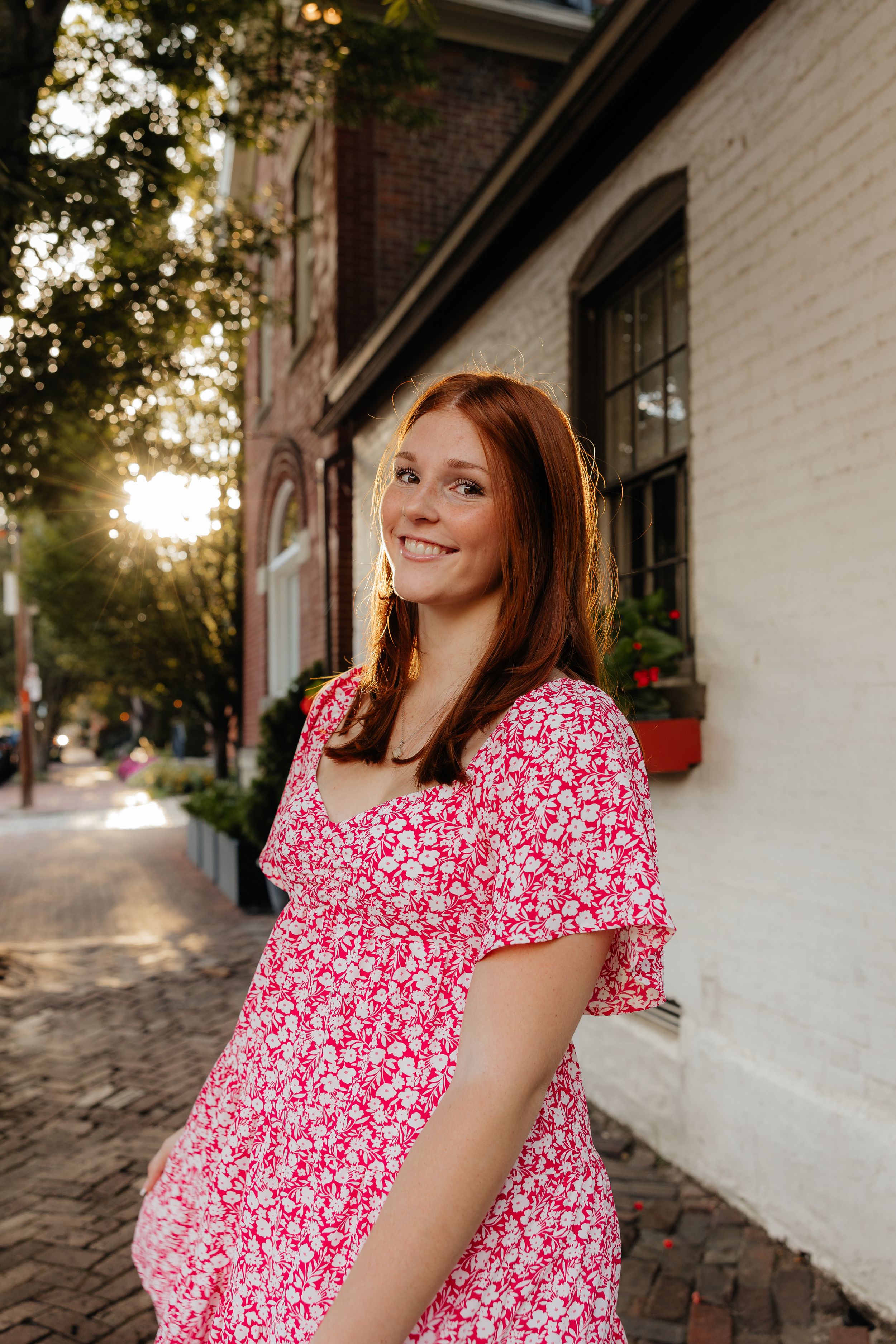 Woman in red floral dress poses along historic brick building sidewalk.