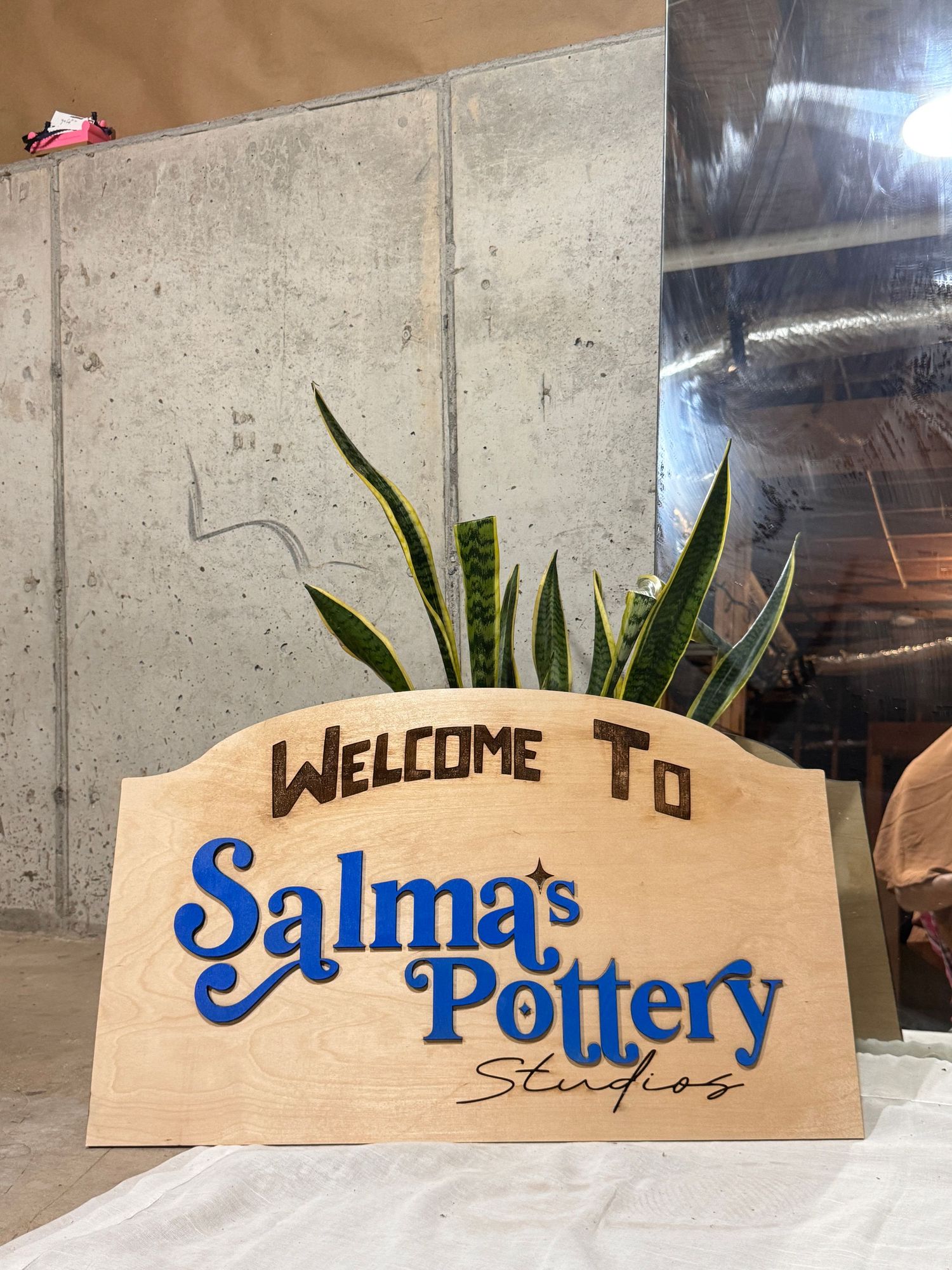 Wooden welcome sign for Salma's Pottery Studios with blue lettering and a snake plant decoration.