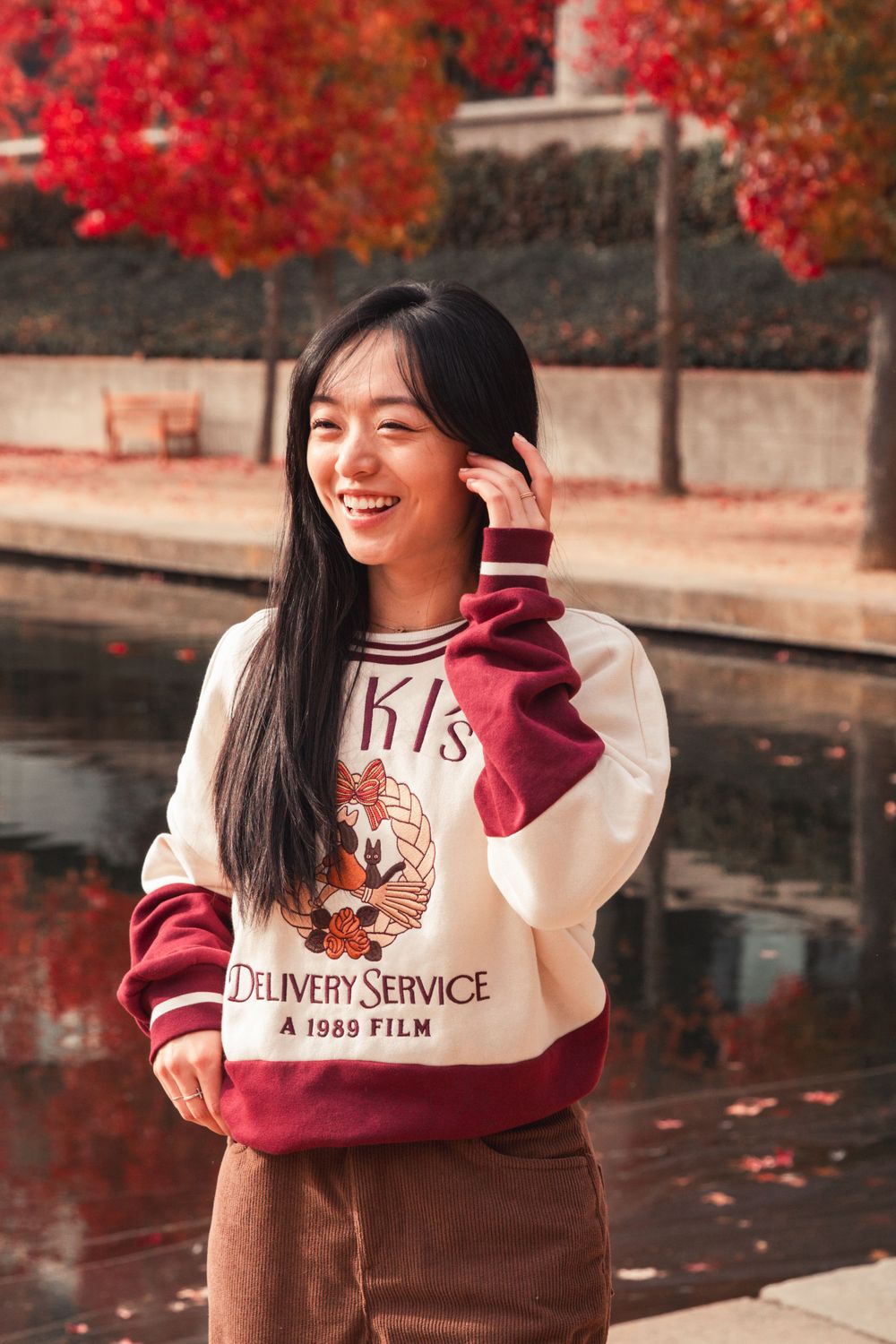 Person in a vintage anime sweatshirt poses by water with red autumn trees in background.