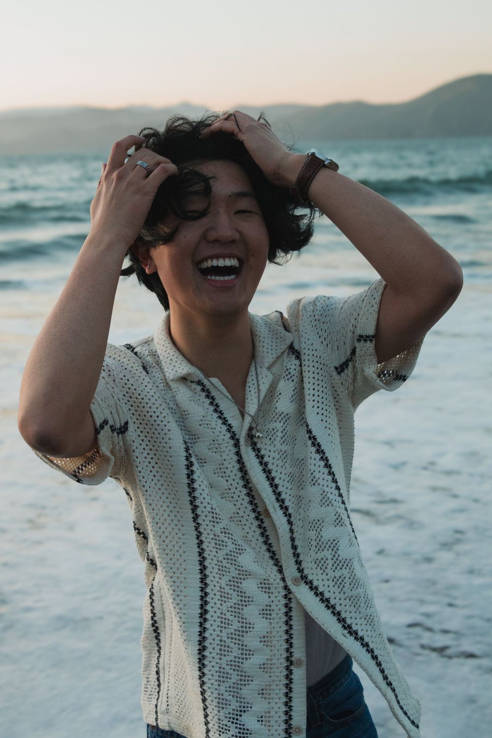 Joyful expression at sunset on a beach wearing a white patterned shirt with waves and mountains in background.
