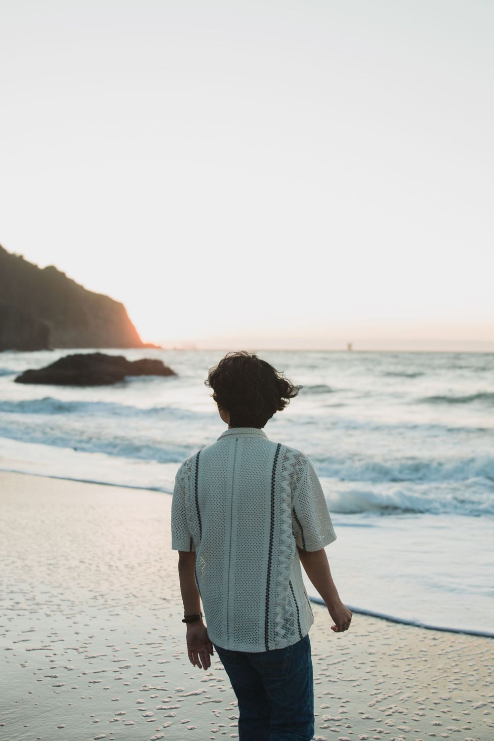 Person in white shirt walking alone on beach at sunset with waves crashing nearby.