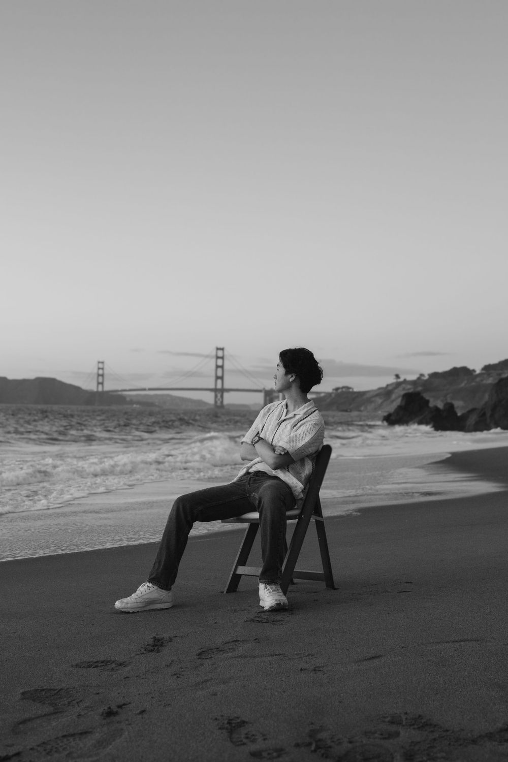 Black and white photo of person sitting on chair at beach with Golden Gate Bridge in distance.
