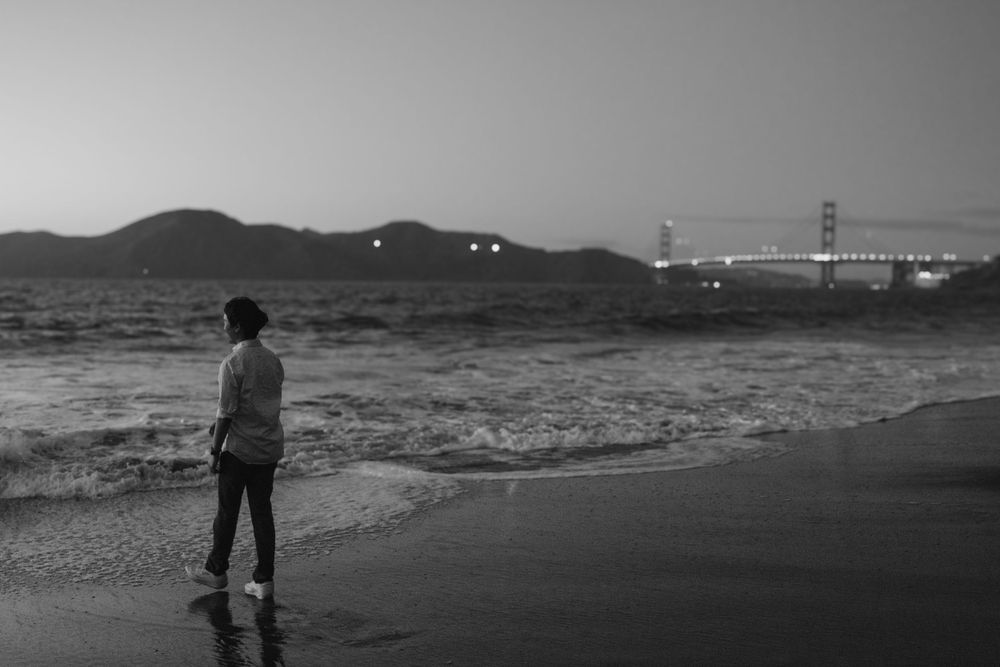 Silhouetted figure standing on beach at dusk with Golden Gate Bridge lights twinkling in distance.