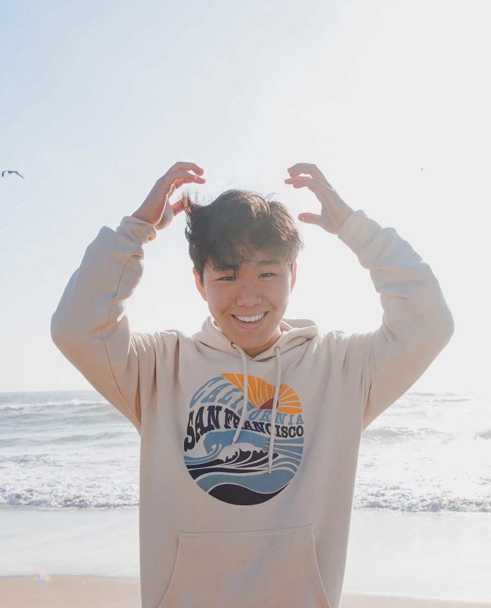 Person in beige hoodie making playful gestures at sunny beach.