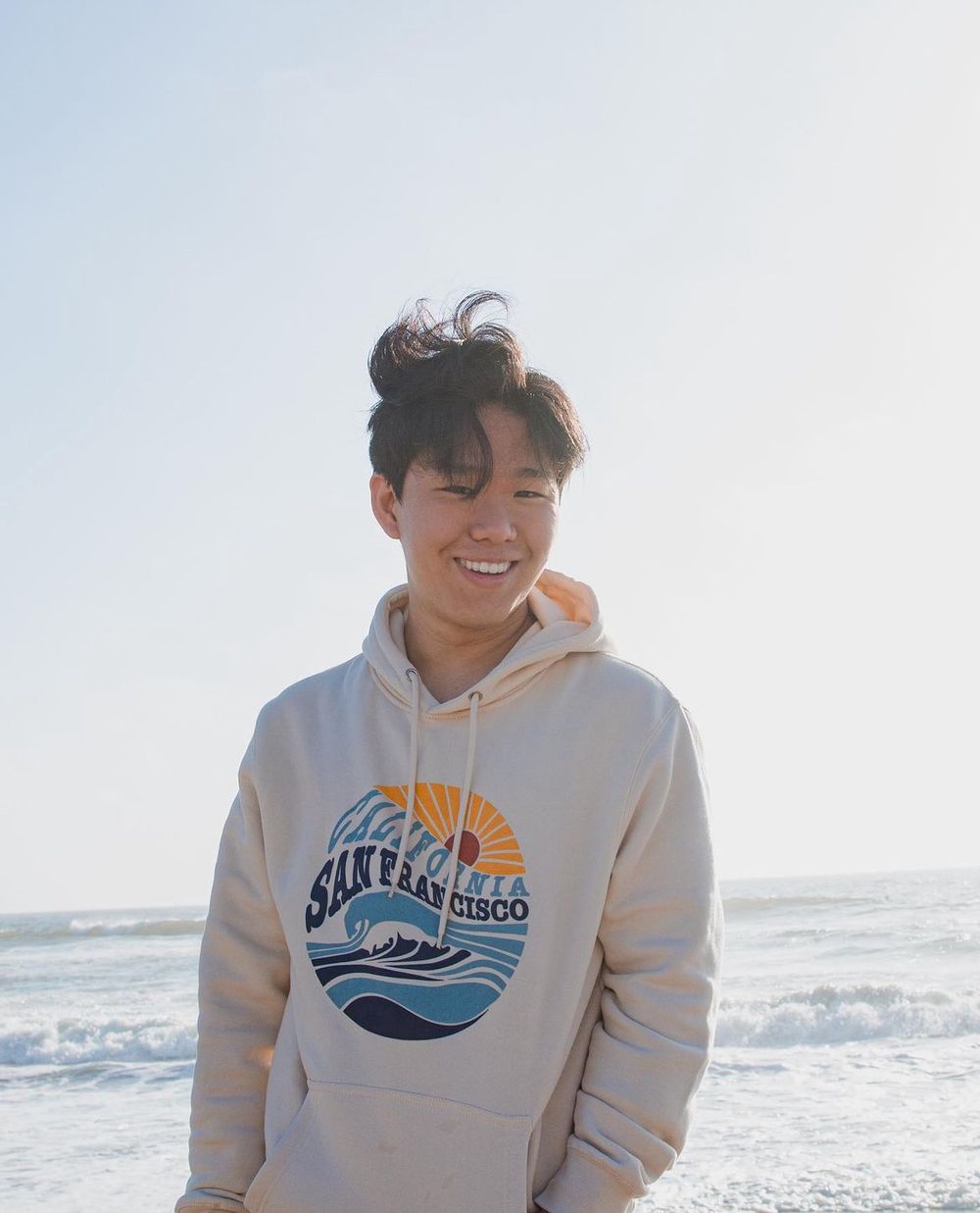 Person wearing cream colored Santa Cruz hoodie smiling at the beach.