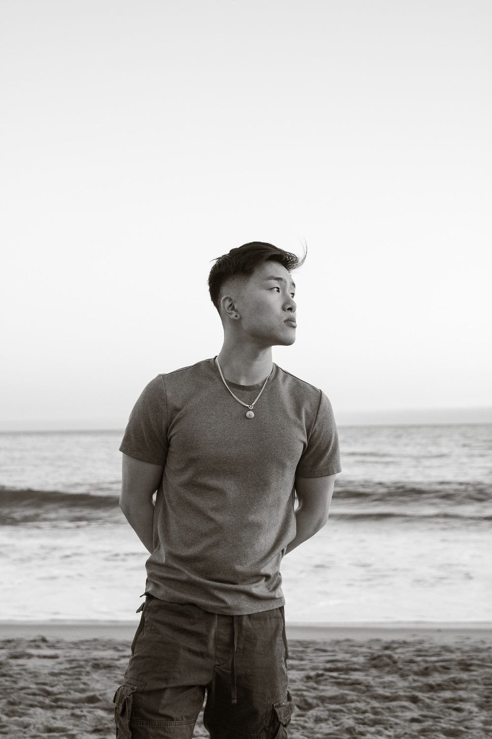 Black and white portrait of a person in a t-shirt standing on a beach at sunset.