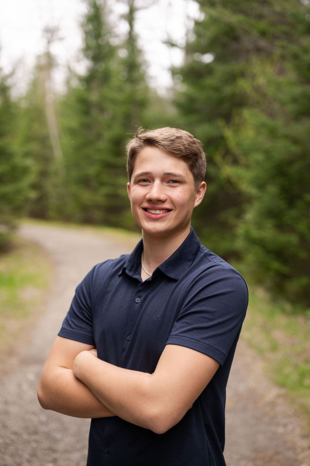 A smiling person wearing a navy polo shirt poses outdoors against a forested background.
