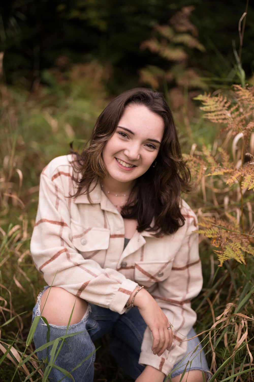 Person in a beige striped shirt and jeans sits casually among autumn ferns in an outdoor nature setting.