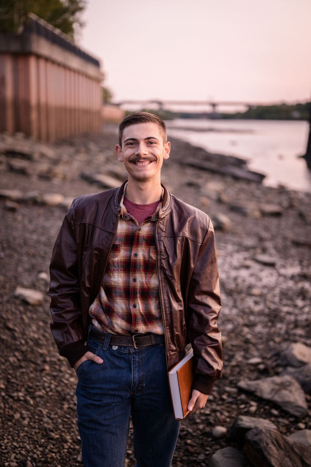 A person in a brown leather jacket and plaid shirt poses casually on a rocky beach at sunset.