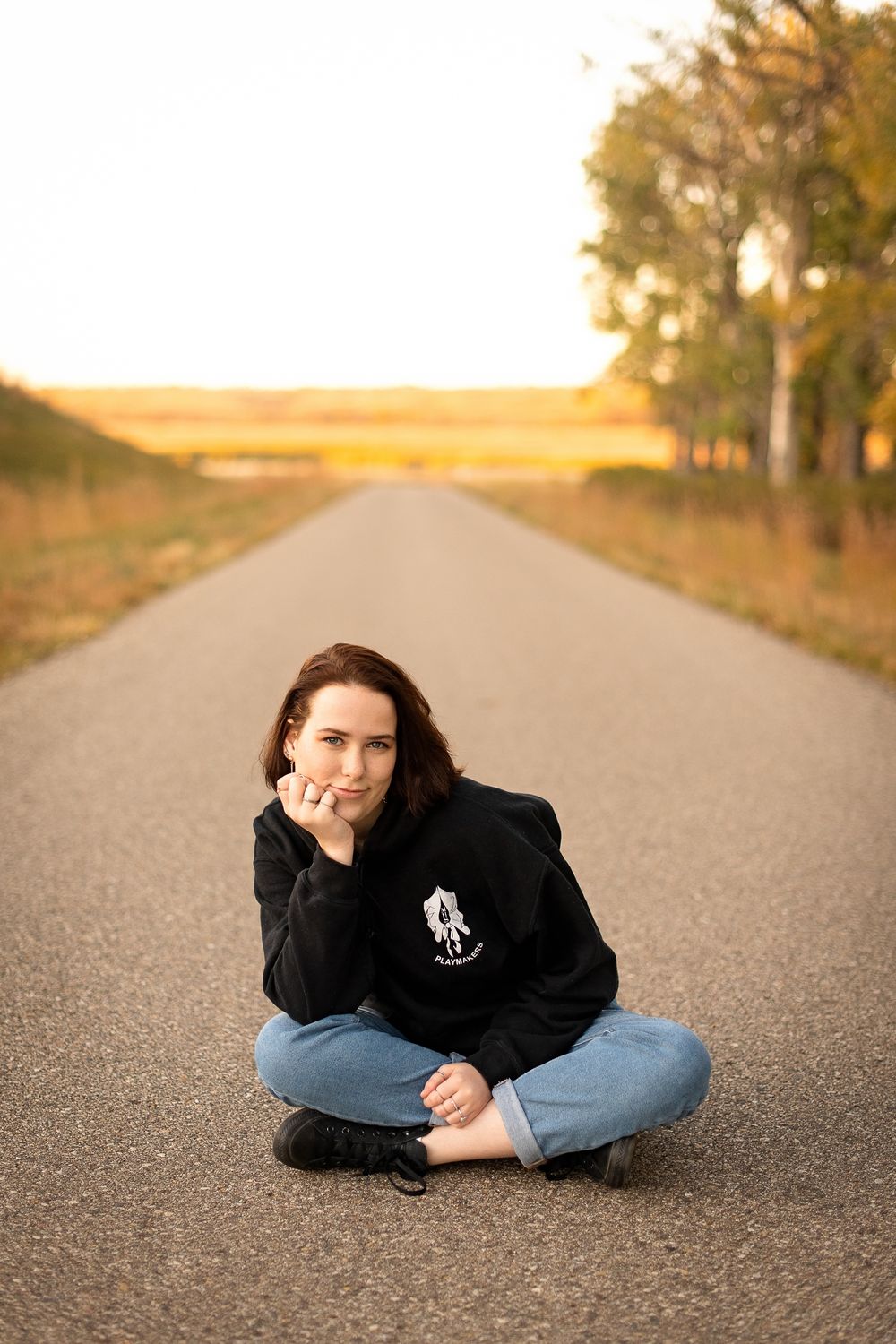 A person sits cross-legged on a rural road at sunset wearing a black hoodie and jeans.