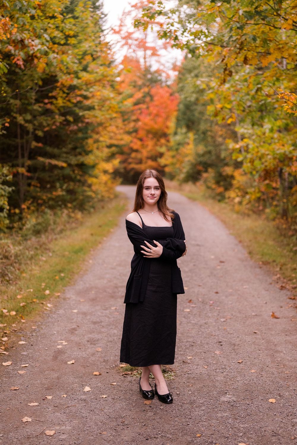 A figure in a black dress poses on a scenic autumn path lined with colorful fall foliage and trees.