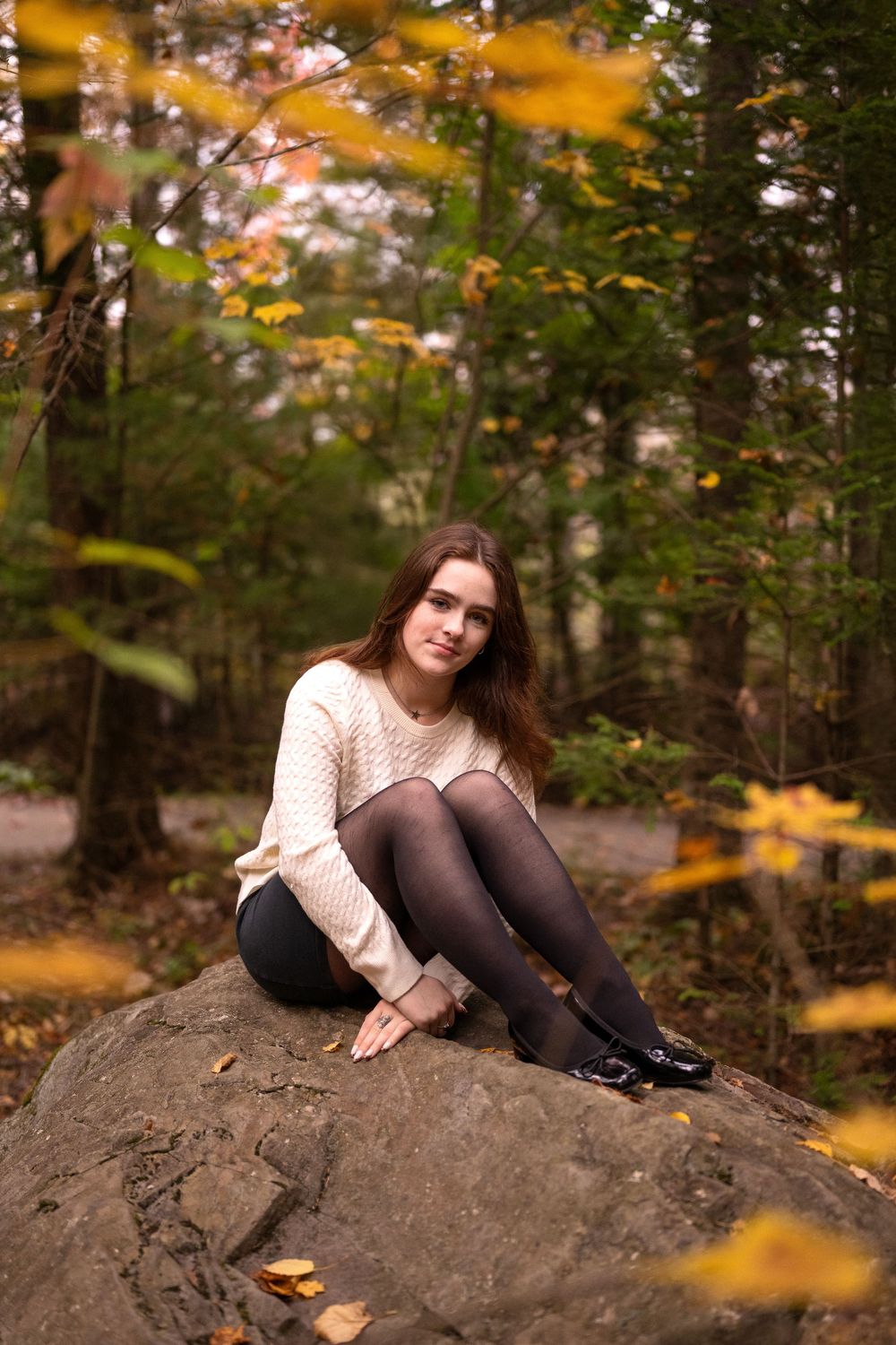A person in a cream sweater and dark tights reclines on a rocky outcrop surrounded by autumn foliage in a forest setting.