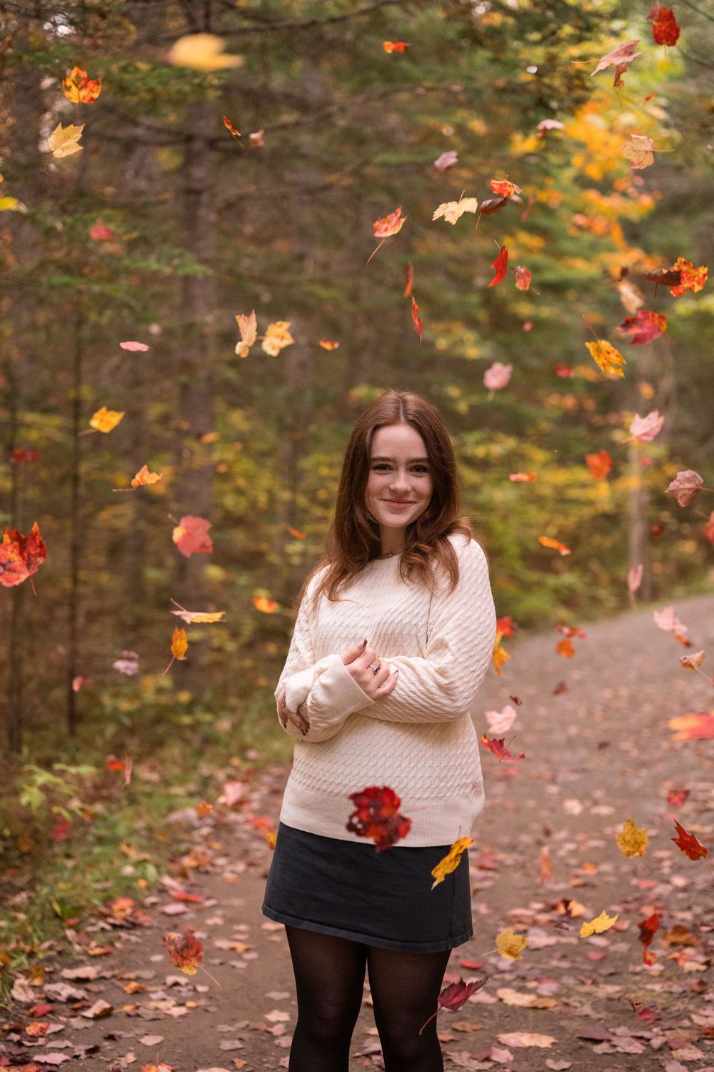 A person in a white sweater stands on a forest path while autumn leaves fall around them in a magical scene.
