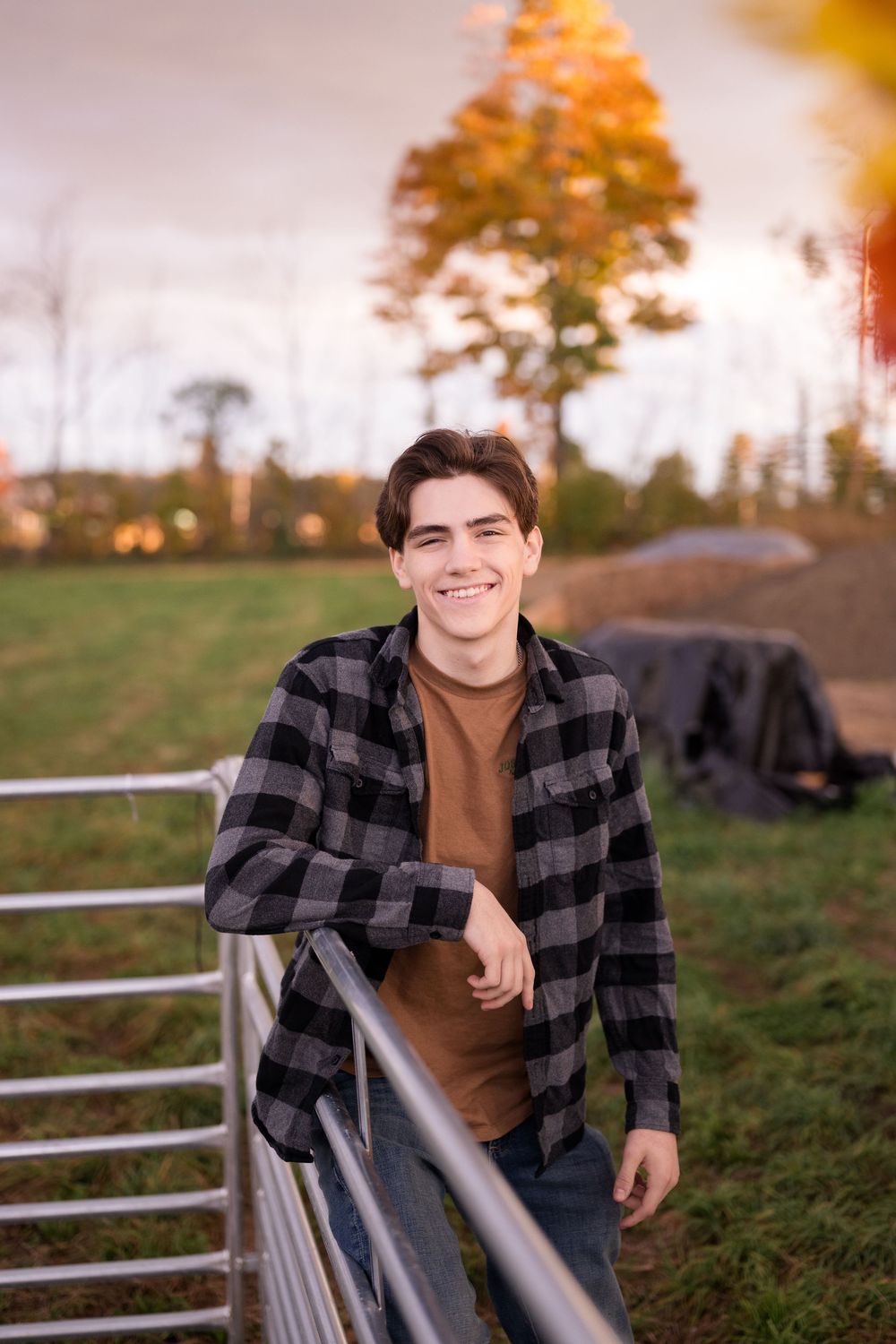 A person in a plaid flannel shirt poses casually against a white fence at sunset in a rural setting.