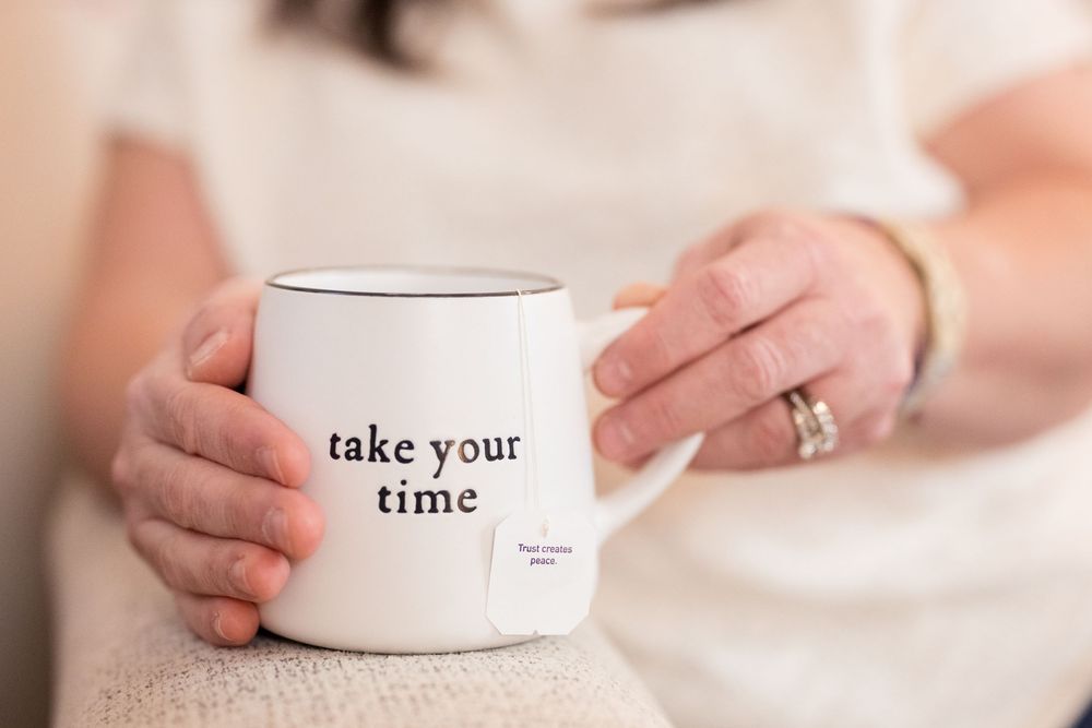 A white ceramic coffee mug with text reading 'take your time' sits on a light neutral surface.