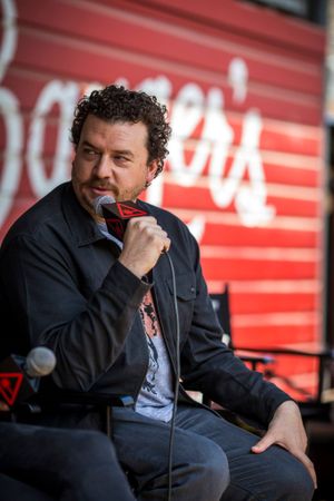 Speaker holding microphone against red wall background during outdoor event.