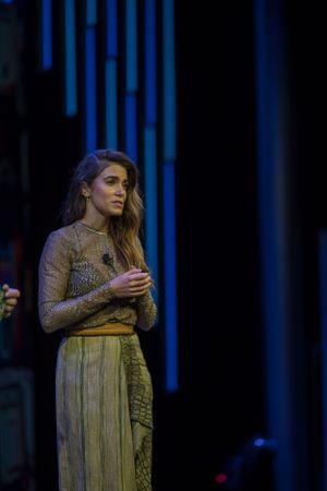 Performance shot of someone in gold dress against dark background with blue stage lighting.