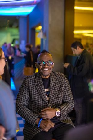 Attendee in patterned blazer and glasses smiles during tech conference with colorful lighting.