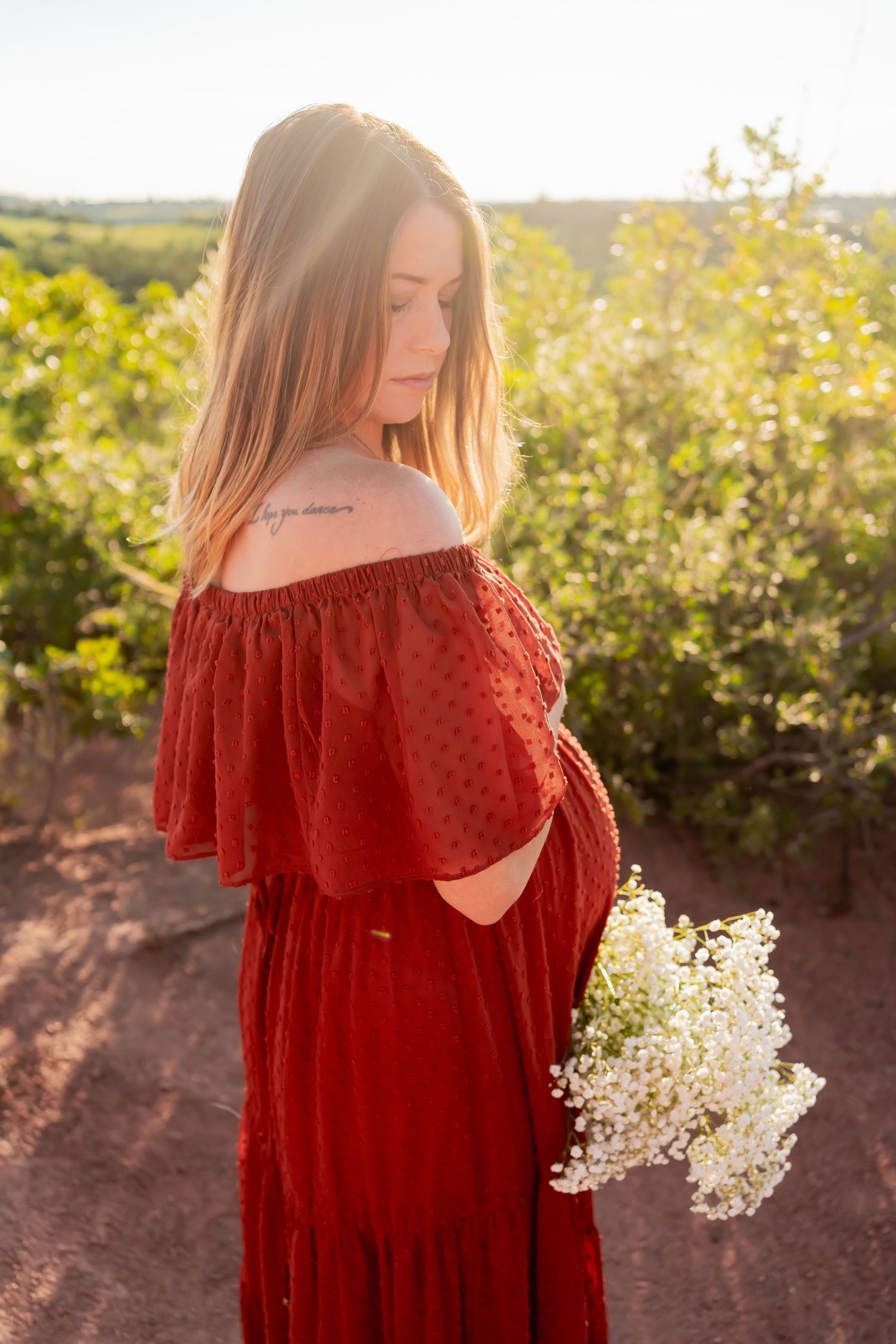 Sunset maternity photo in red off-shoulder dress holding white flowers.