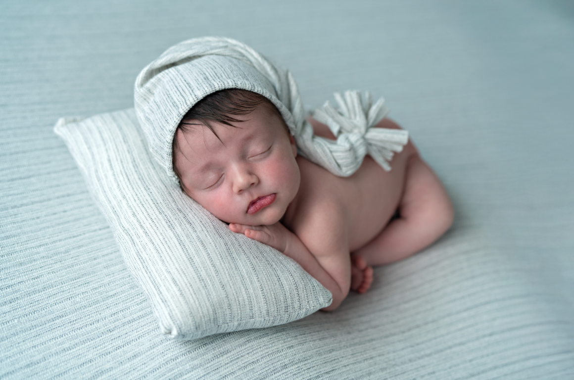 Sleeping newborn baby wearing a white knit bonnet resting on a small pillow during a studio photography session. Embracing Life’s Beauty