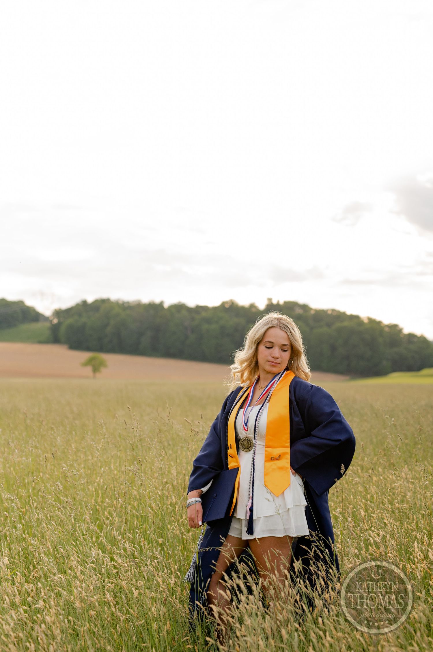 Graduate in navy gown and gold stole standing in tall grass with rolling hills behind.