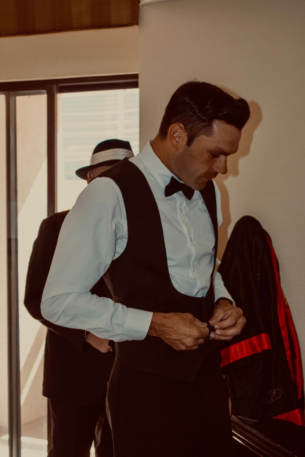 Groom in black vest and bow tie buttoning up while getting ready for wedding day.