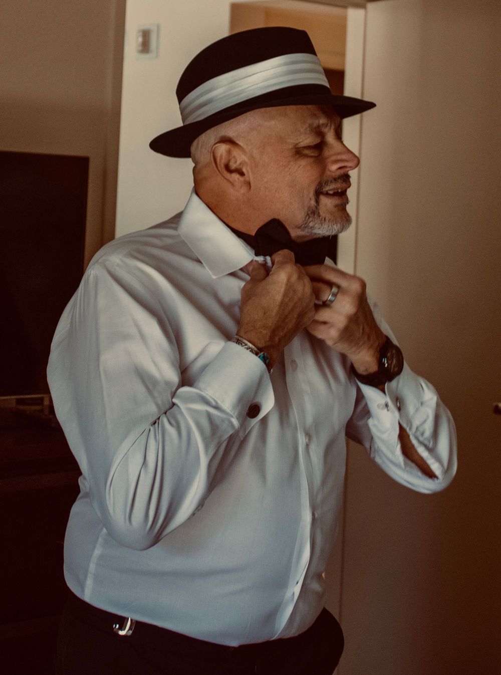 Man in white shirt and black fedora hat adjusting bow tie while getting dressed for formal event.