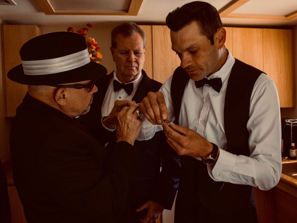 Three men in formal tuxedos and bow ties examine a small object together indoors.
