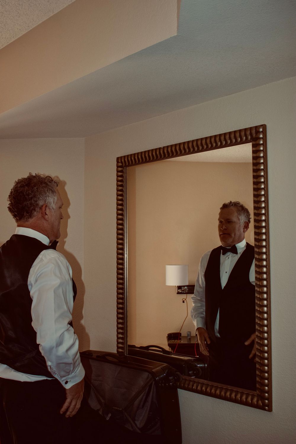 Man in tuxedo vest and bow tie checking his appearance in an ornate gold-framed mirror in a hotel room.