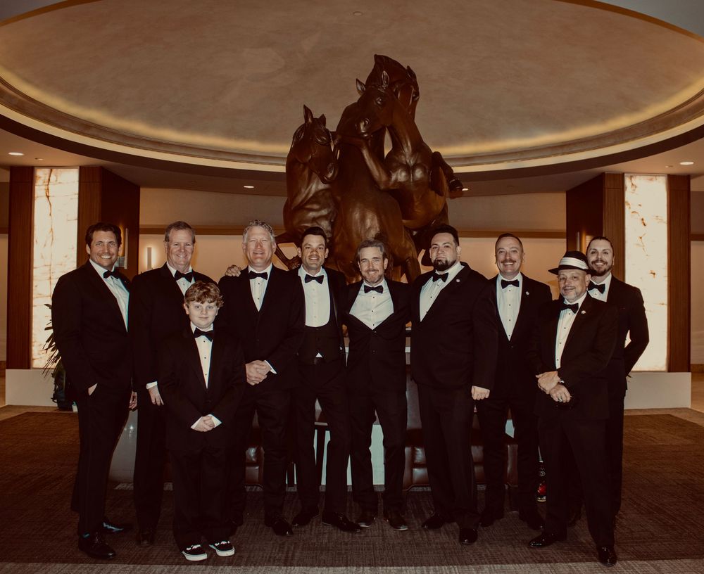 Group of men in tuxedos posing formally in front of a bronze horse sculpture in an elegant venue.