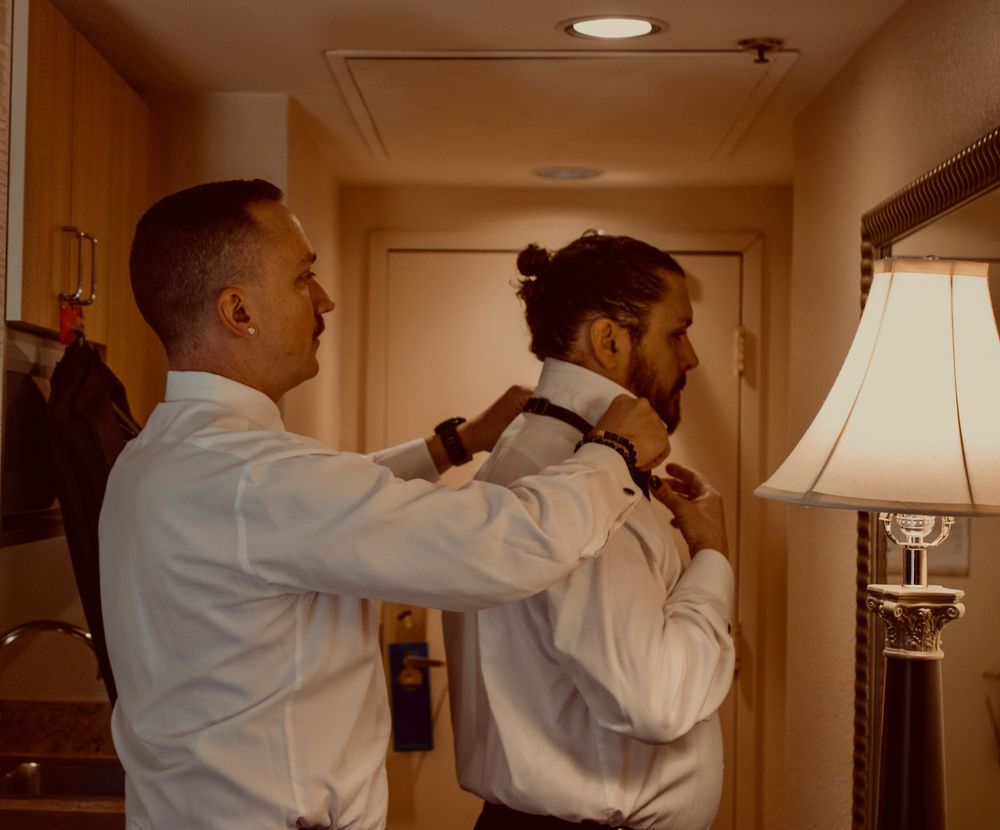 Two men in white dress shirts prepare for a wedding, one helping the other adjust his tie in a hotel room.