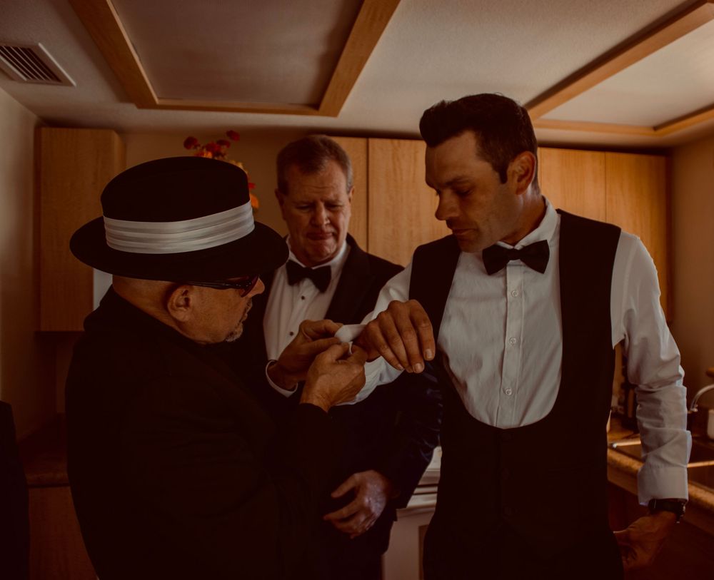 Three men in formal attire adjust cufflinks before a wedding ceremony indoors.