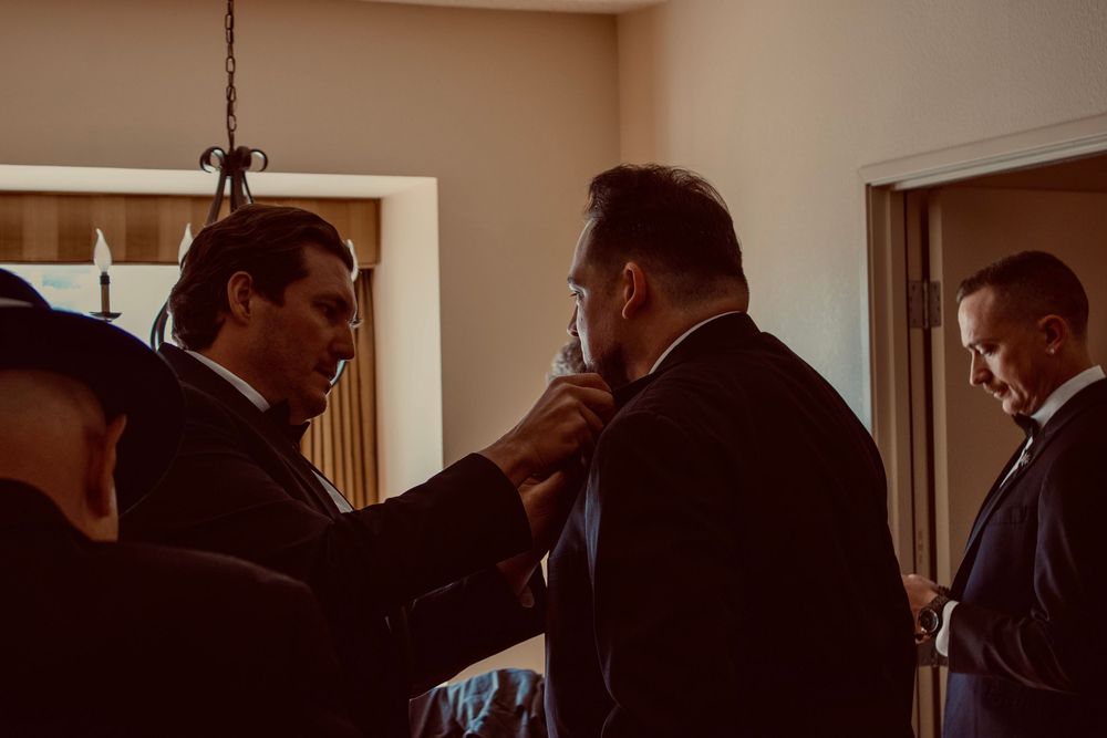Groomsmen helping each other adjust ties while preparing for a wedding ceremony indoors.