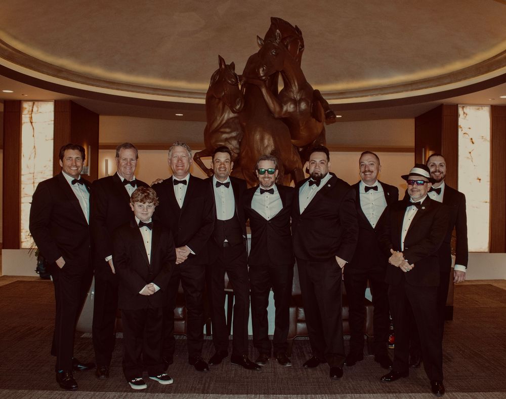 Group of groomsmen in matching black tuxedos and bow ties posing before a bronze horse sculpture.